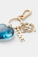 Llavero metálico dorado con gancho mosquetón y argolla, decorado con dijes colgantes: un trébol de cuatro hojas con pedrería, un pequeño animal alado, una placa con la palabra 'LOVE' y un gran corazón azul translúcido de acrílico.