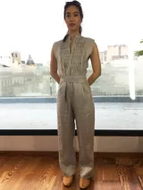 Mono largo de lino color beige, con detalle plisado en el frente, escote en V, sin mangas, lazo en la cintura, cierre invisible delantero y bolsillos laterales.