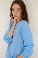 Blusa celeste de manga larga con delicados bordados florales en tono más claro en la zona del pecho y mangas. Presenta escote en V y puños elásticos abotonados.