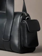 Cartera estructurada Calvin Klein de piel sintética negra, con diseño de asas superiores y bolsillos laterales. Cuenta con cierre de cremallera y logo metálico grabado en la parte delantera.