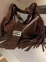 Cartera tipo hobo de gamuza color marrón, con diseño de media luna y flecos laterales. Presenta una manija trenzada, cierre metálico superior y una solapa frontal con hebilla metálica plateada con la letra S y el nombre Sofia grabado.