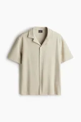 Camisa de manga corta color beige con textura de espiga, cuello abierto, tapeta lisa y bajo recto con abertura en los laterales.