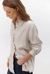 Camisa beige de manga larga con botones marrones y cuello clásico. Presenta un detalle de costura dorada a lo largo de la botonera.