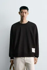 Sudadera negra de corte relaxed fit, confeccionada en tejido técnico interlock de algodón con elasticidad. Presenta cuello redondo, manga larga y una etiqueta aplicada en el bajo. Los acabados son en rib.