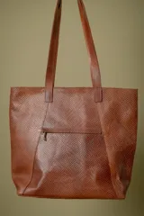 Bolso tipo shopper de cuero color suela con textura grabada, detalle de costuras laterales trenzadas y placa metálica con logo de la marca. Cuenta con doble asa larga para colgar al hombro, cierre principal y múltiples bolsillos internos y externos.