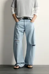 Jeans de corte barrel fit confeccionados en denim de algodón rígido, con diseño de cinco bolsillos y cierre frontal con botones. Presentan un lavado claro con efecto de roturas en las perneras.