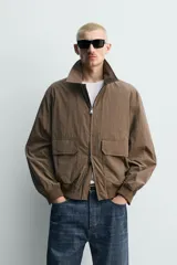 Cazadora regular fit de tejido técnico color marrón, con cuello solapa, manga larga y cierre frontal de cremallera. Presenta bolsillos de plastrón con solapa en la cadera y acabados con rib elástico.