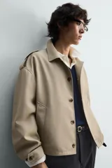 Cazadora color beige, estilo cropped, con cuello de solapa, manga larga con puños ajustables y trabillas en los hombros. Presenta bolsillos de vivo en la cadera y cierre frontal de botonadura.