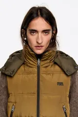 Chaleco acolchado de largo medio y corte en línea A, confeccionado en tejido técnico color beige con detalles en negro. Presenta cuello subido, capucha desmontable, cierre frontal de cremallera, bolsillos laterales con cremallera, logo bordado en el pecho y aberturas laterales con cremallera.