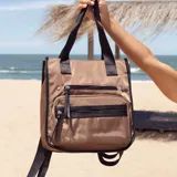 Mochila y cartera híbrida confeccionada en nylon de color beige con detalles y correas en negro. Posee dos asas superiores para llevar al hombro y correas traseras ajustables para usar como mochila. Presenta dos bolsillos frontales con cierre de cremallera y tiradores largos.