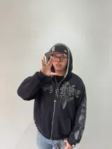 Campera reversible con cierre frontal, diseño oversize. Un lado es de algodón negro con estampados y bordados gráficos, mientras que el otro lado es de tela polar gris de pelo largo. Cuenta con capucha, puños y ruedo acanalados.