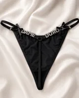Tanga de hilo negra con tiras regulables, moños laterales y detalle de la frase "Dame Duro" en pedrería brillante en la parte frontal.