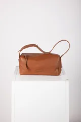 Cartera tipo baguette confeccionada en cuero de color tostado, con silueta alargada y cierre superior en diagonal. Cuenta con una correa para el hombro con detalles trenzados y un aplique con logo grabado.