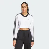 Blusa cropped de manga larga Adidas Adicolor en color blanco, con cuello en V y puños acanalados en negro. Presenta las tres tiras clásicas de la marca en color negro a lo largo de las mangas y el logo del trifolio bordado en el pecho.