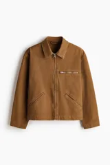 Campera de algodón color marrón con cuello camisero, cierre frontal, bolsillo superior con cierre y bolsillos laterales ribeteados.