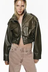 Cazadora corta de efecto piel en color verde militar, con cuello subido, cierre frontal con cremallera y botones a presión ocultos. Presenta mangas largas con trabillas en los hombros y bolsillos frontales con solapa. El diseño incluye un dobladillo inferior elástico acanalado.