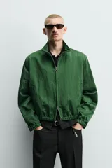 Campera verde oscuro de corte cropped y tejido técnico, con cuello alto, cierre frontal de cremallera con tirador circular, bolsillos ribeteados y puños y dobladillo elásticos.