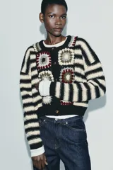 Chaqueta de punto con cuello redondo y manga larga, que combina un diseño de rayas horizontales en blanco y negro con paneles frontales decorados con motivos de crochet en tonos tierra y granate. Presenta cierre frontal con botones.