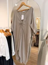 Vestido midi de lino color beige, con diseño oversize, escote en V y detalle de nudo drapeado en el frente. Posee mangas cortas amplias con dobladillo y corte asimétrico, siendo más largo en la parte trasera.