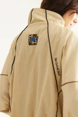Campera rompevientos beige de nylon con diseño oversize, cuello alto, cierre frontal negro y detalles de costuras y ribetes en contraste.
