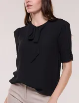 Blusa blanca de corte recto y tejido liviano de gasa con forro interno delantero. Presenta escote en V con cuello redondo y lazo central para anudar, mangas cortas con hilera de botones forrados en los laterales y ruedo ligeramente redondeado.