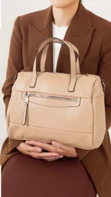 Cartera de mano y hombro color beige, de material símil cuero, con forma estructurada y redondeada. Posee dos asas cortas superiores y una correa larga ajustable y desmontable. Cuenta con un bolsillo exterior con cierre en la parte frontal.