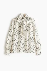 Blusa de gasa color beige con estampado de lunares negros, cuello alto con moña integrada y mangas largas abullonadas con puños ajustados.