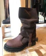 Bota de caña media con diseño slouchy, confeccionada en una combinación de cuero liso y gamuza en tonos marrones. Presenta una suela track robusta de goma negra y una tira decorativa con hebilla metálica en el lateral.