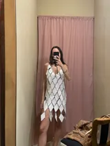 Vestido corto blanco, tipo túnica, confeccionado artesanalmente con cientos de rombos de cuero genuino perforados y unidos entre sí mediante engarces, creando un efecto de malla con aberturas. Presenta escote en V profundo y tirantes finos.