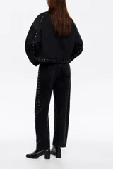 Chaqueta de jean negra de corte oversize, con cierre frontal de botones metálicos y detalle de tachas plateadas distribuidas en el frente y los hombros.