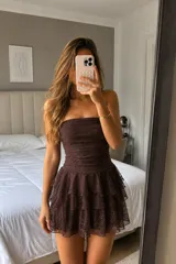 Vestido corto strapless de color marrón con acabado brillante. Presenta un diseño ajustado en el torso con frunces y una falda con volados superpuestos de tul con destellos.