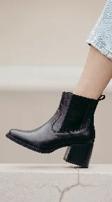 Bota tipo Chelsea de cuero negro, con paneles laterales elásticos y taco cuadrado de 6 cm con acabado de madera. Diseño clásico con puntera redondeada y tirador trasero.