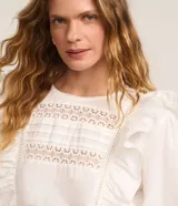 Blusa blanca de tricolina con diseño romántico, cuello redondo, mangas largas abullonadas y detalles de volados verticales en el frente. Presenta aplicaciones de encaje tipo entremedios en el pecho y en las mangas.