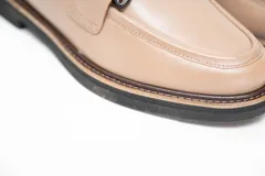 Mocasines de cuero vacuno color visón, con costuras visibles en la parte superior y un adorno metálico negro en el empeine. Poseen suela baja y diseño sin cordones.