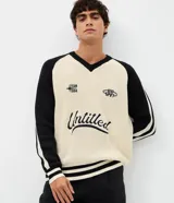Suéter masculino de punto en modelo boxy, con cuello en V, manga larga y diseño bicolor en beige y negro. Presenta inscripciones bordadas en el pecho, incluyendo la palabra 'Untitled' en tipografía cursiva, y detalles de rayas en los puños, las mangas y el ruedo.