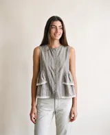 Blusa sin mangas de diseño fluido, confeccionada en tejido gris con rayas verticales finas. Presenta cuello redondo, cierre frontal con botones y detalles de volados escalonados en los laterales con terminaciones en puntilla blanca.