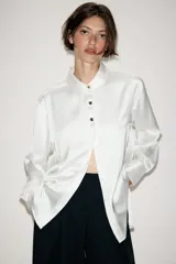 Camisa de satén color blanco, de corte clásico con cuello solapa, manga larga con puños y cierre frontal con botones dorados.