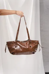 Cartera tipo shopper de cuero sintético marrón, con diseño alargado y flexible. Cuenta con asas largas de hombro ajustables con hebillas, detalles de cordones laterales con ojales metálicos y una pequeña placa con el logo de la marca en la parte frontal.