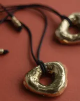 Collar con dije de forma irregular en tonos dorados y plateados, suspendido de un cordón doble de algodón negro.