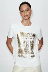 Camiseta de algodón de corte regular con cuello redondo y manga corta. Presenta un estampado frontal metalizado en tono dorado con diseño gráfico.