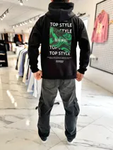 Canguro oversize negro con capucha y bolsillo tipo canguro. Presenta un estampado gráfico en la espalda con la inscripción "TOP STYLE", un rectángulo con efecto marmolado en verde y negro con la palabra "WORLDWIDE", y texto descriptivo adicional con un código de barras.