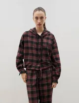 Camisa de manga larga con capucha y cordones ajustables, confeccionada en tejido liviano con estampado a cuadros en tonos bordó y negro. Presenta cierre frontal con botones y un lazo regulable en la cintura.