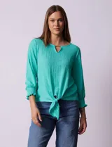 Blusa blanca de tejido texturado con cuello redondo, escote en V con aplique metálico y mangas 3/4 con puños elásticos. El ruedo se ata con un nudo.