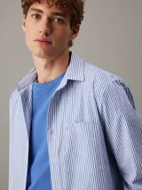 Camisa de manga larga Calvin Klein confeccionada en popelina de algodón elástico con diseño de rayas verticales. Presenta cuello en punta, cierre frontal de botones, bolsillo de parche en el pecho con logo bordado y bajo redondeado.