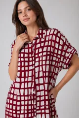Vestido camisero de corte holgado y silueta relajada, con cuello clásico, cierre frontal de botones y mangas cortas con dobladillo. Presenta un estampado de cuadros irregulares en color bordó sobre fondo blanco.