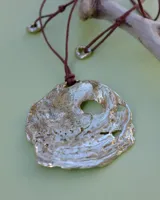 Collar artesanal con dije central de metal con textura orgánica irregular que evoca una ostra. El dije está montado sobre un cordón de algodón natural, permitiendo un uso versátil y regulable.