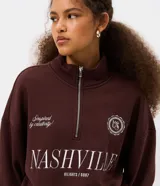 Buzo de algodón y poliéster color marrón, con cuello alto y cierre frontal parcial. Presenta diseño de hombros caídos, puños y ruedo acanalados, y estampado tipográfico frontal con la palabra Nashville.