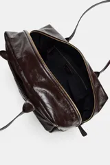 Bolso tipo bowling de gran tamaño confeccionado en piel con acabado brillante. Presenta doble asa de hombro, cierre principal mediante cremallera, bolsillos exteriores en la parte delantera y trasera, y un bolsillo interior con cierre.