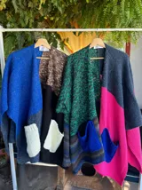 Ruana abierta de tejido de punto con diseño de bloques de color y bolsillos frontales aplicados. Presenta una textura variada y un corte holgado, ideal para superponer sobre otras prendas.