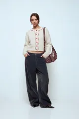 Cardigan de punto color beige, de corte cropped y manga larga. Presenta un diseño con cuello redondo y una hilera de botones frontales en color rojo, con detalles de costuras decorativas alrededor del cuello y la botonera.